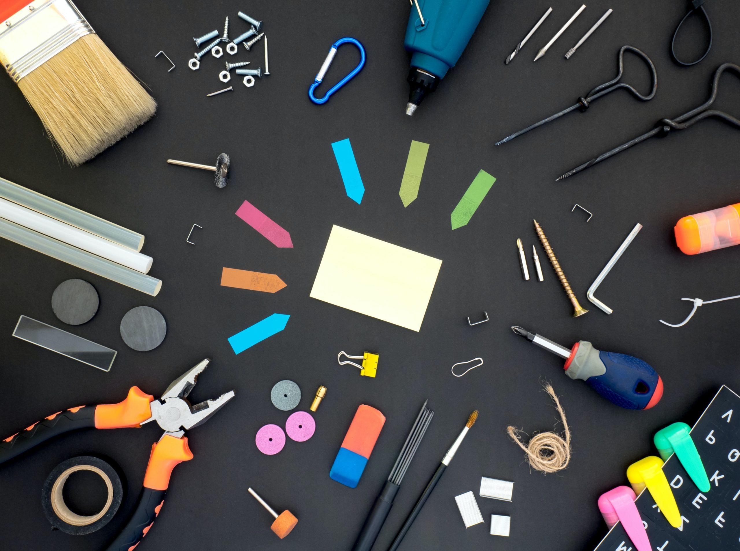 An assortment of tools laying on a black surface including post-its, screwdrivers, wrenches, tape, paintbrushes, highlighters, glue sticks, staples, magnets, and paperclips.